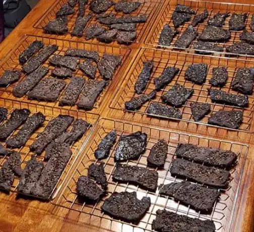 Six trays of smoked meat are resting on top of a wooden table to review the Bradley Electric Smoker by Pioneer Smoke Houses.